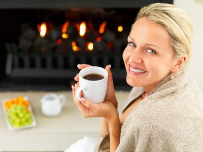 A woman wrapped in a towel holds a cup, sitting by a fireplace. A plate of fruit is in the background.