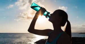 Silhouette of a person drinking from a water bottle by the sea with the sun setting in the background.