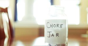 A glass jar labeled "Chore Jar" sits on a wooden table in a softly lit room.