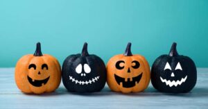 Four decorative pumpkins with painted faces, two orange and two black, are arranged in a row on a light blue surface against a teal background.