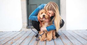 A woman in a denim jacket hugs a fluffy, brown dog sitting on a wooden porch. The dog is on a leash and facing the camera.