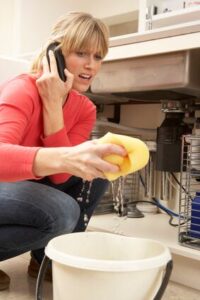 A woman is crouching under a sink, holding a yellow sponge with water dripping into a bucket, while talking on a phone.