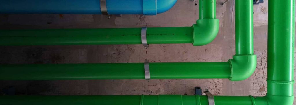Blue and green parallel pipes with metal clamps run horizontally along a rough concrete wall.