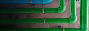 Blue and green parallel pipes with metal clamps run horizontally along a rough concrete wall.