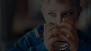 A young child in a blue sweatshirt drinks from a glass, looking to the side.