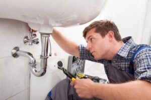 A plumber in a plaid shirt uses tools to fix a pipe under a bathroom sink.
