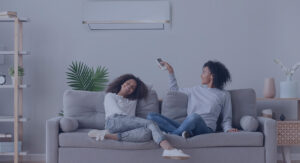 Two people sitting on a gray couch; one holds a remote control pointed at the wall-mounted air conditioner above. Shelves and plants decorate the room.
