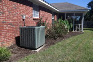 Air conditioning unit outside a brick house, next to a garden with shrubs and flowers. There is a screened porch attached to the house. Grass lawn in the foreground.