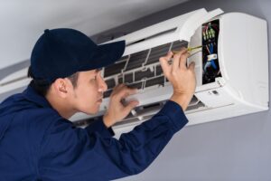 Technician in blue uniform and hat repairs an open wall-mounted air conditioning unit using a screwdriver.