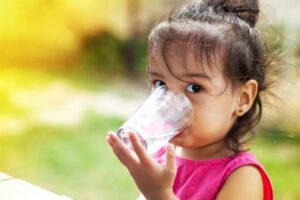 A young girl in a pink shirt drinks water from a clear glass, looking at the camera.