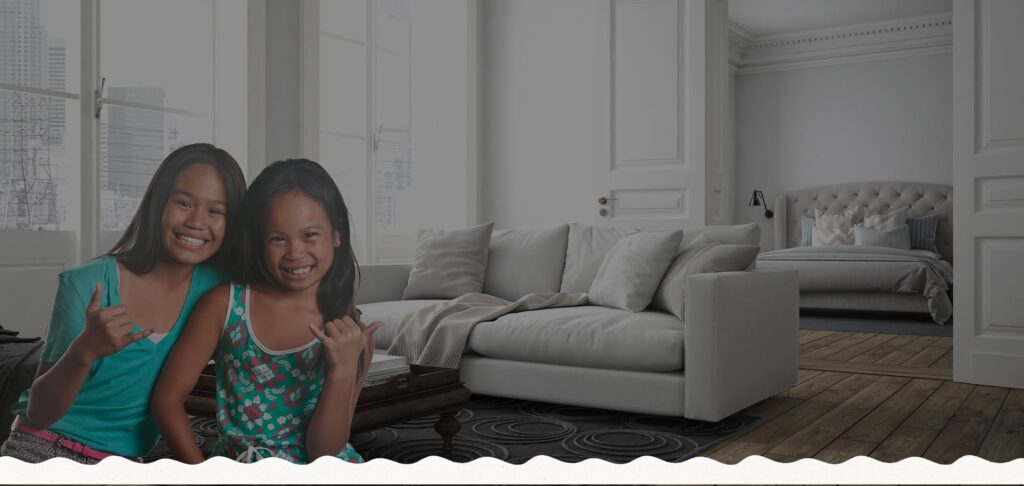 Two smiling children in colorful clothing gesture with their hands in a modern living room with a sofa and a view of a bed in an adjoining room.