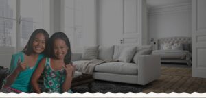 Two smiling children in colorful clothing gesture with their hands in a modern living room with a sofa and a view of a bed in an adjoining room.