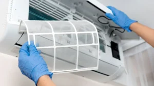 Hands wearing blue gloves replacing a filter in an open wall-mounted air conditioning unit.