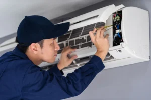 Technician in blue uniform and cap repairing a wall-mounted air conditioning unit with a screwdriver.