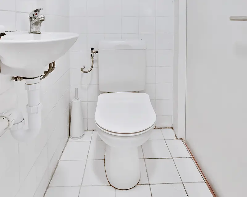 A white bathroom with a toilet, sink, and tiled floor and walls. A toilet brush is placed beside the toilet.