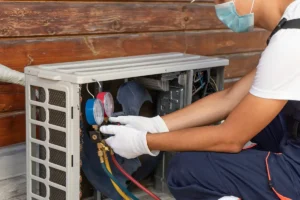 A technician wearing a mask and gloves checks the pressure gauges on an air conditioning unit mounted outdoors on a wooden wall.
