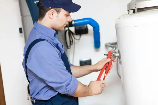 A person in a cap and overalls uses a wrench to adjust a valve on a large white tank in a utility room.